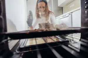 A woman opening the oven and discovering she burnt food inside. 